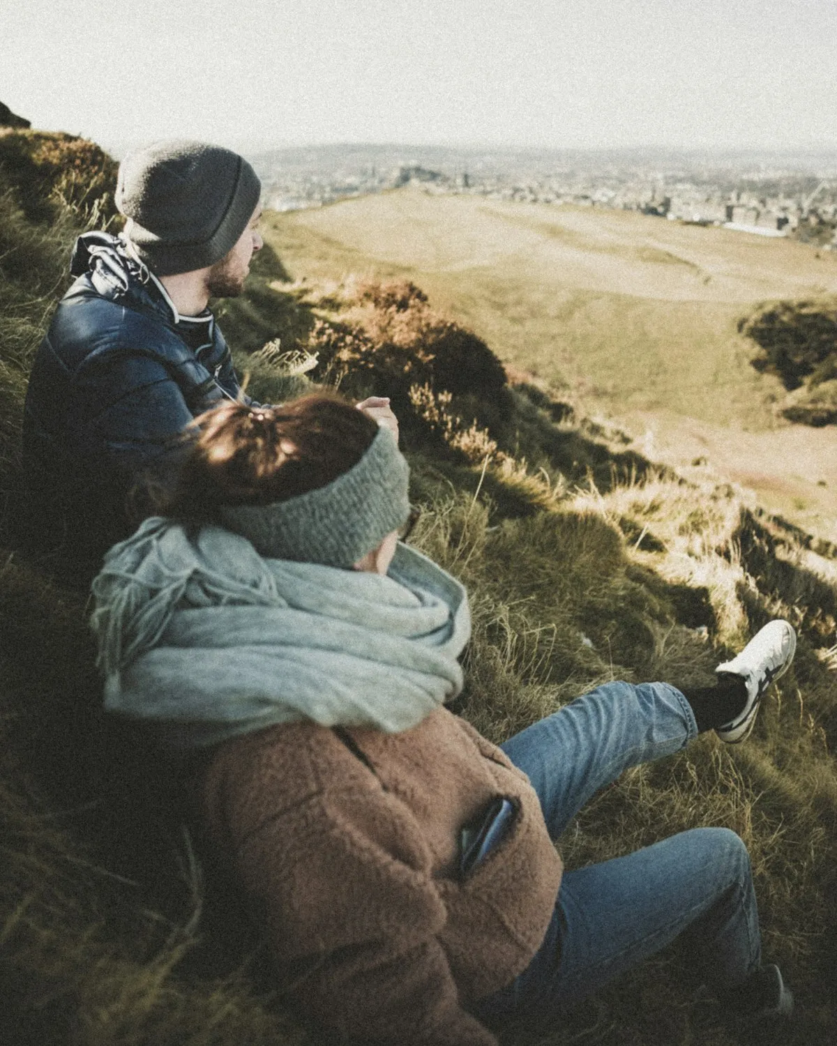 Two people wrapped up warm on a grassy hillside, looking out over Edinburgh