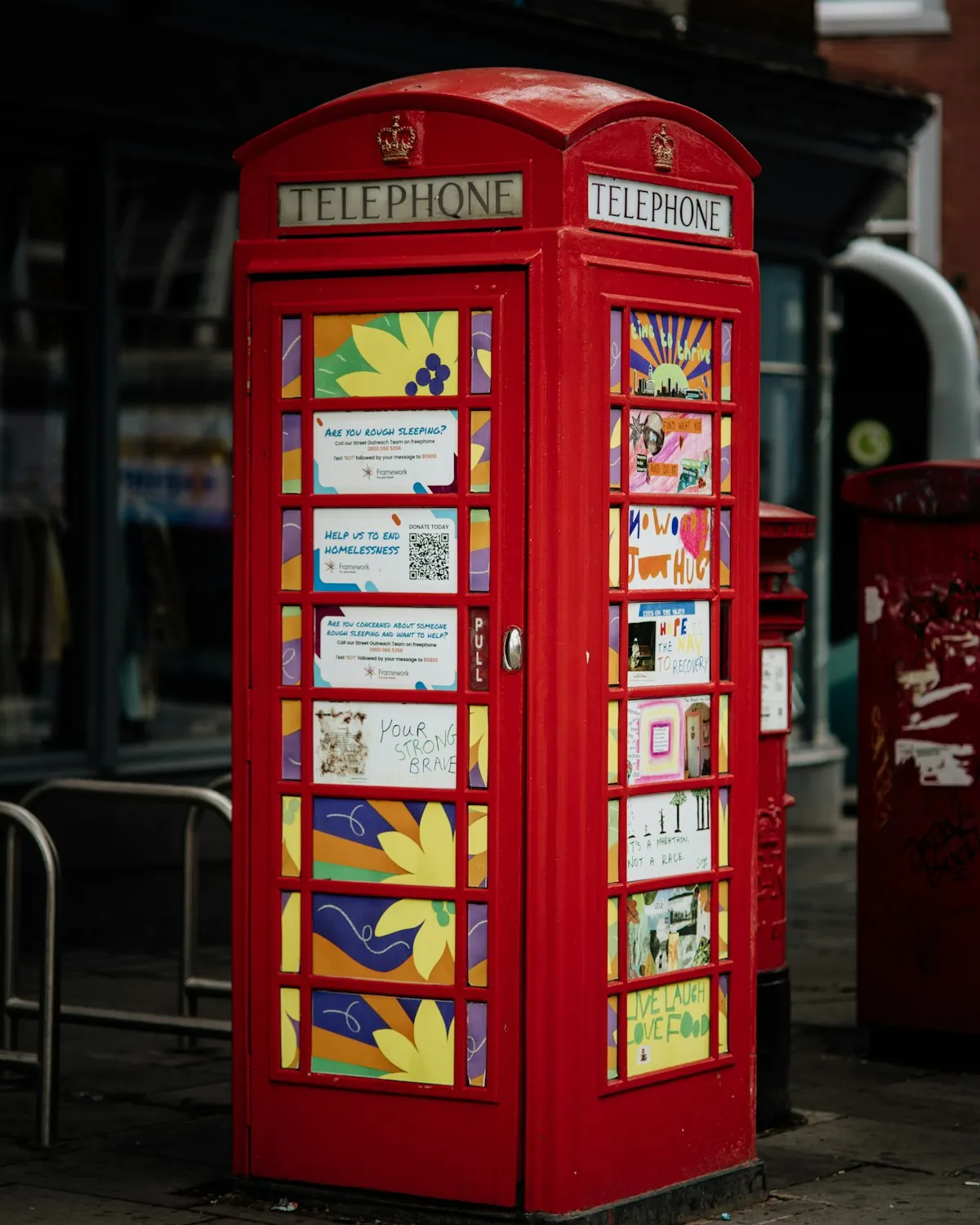 A red telephone box on a British street, decorated with hopeful artwork and posters offering help to people sleeping rough