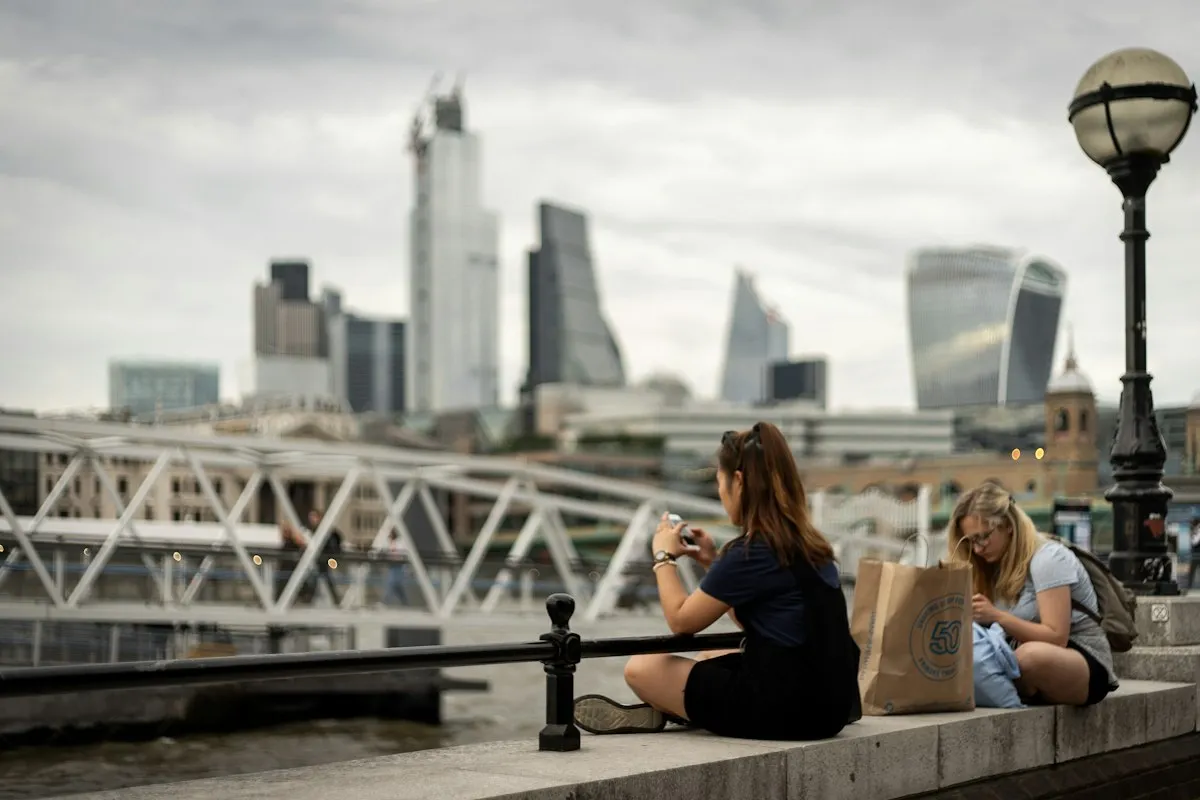 Two women sitting by the Thames with the City of London skyline behind them