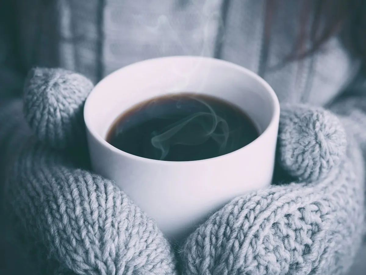 Hands in knitted mittens holding a steaming mug of coffee